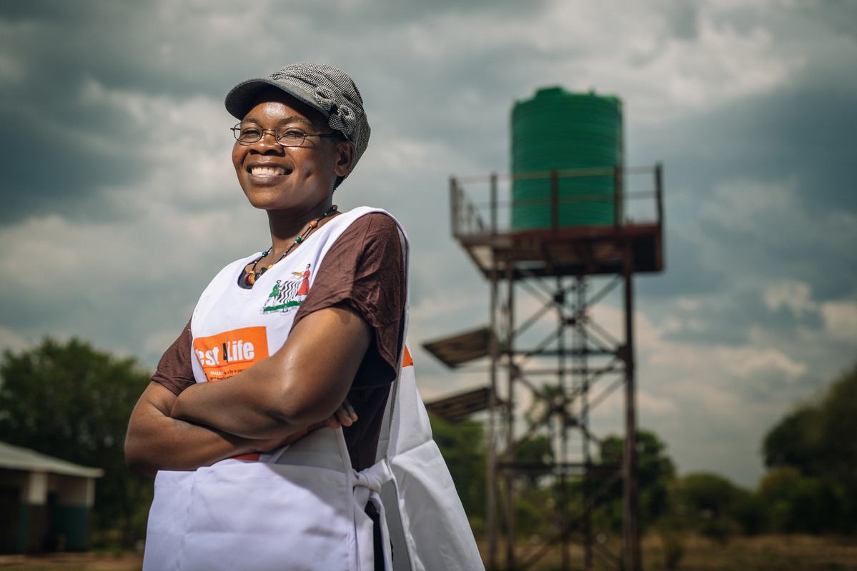 17486 Precious Namakau, community health worker, 38 years old, 3 children, wearing cap and glasses, test and treat apron, stands outside with the Mandia community health post's water tower in the background. Photo: PATH/Gabe Bienczycki.