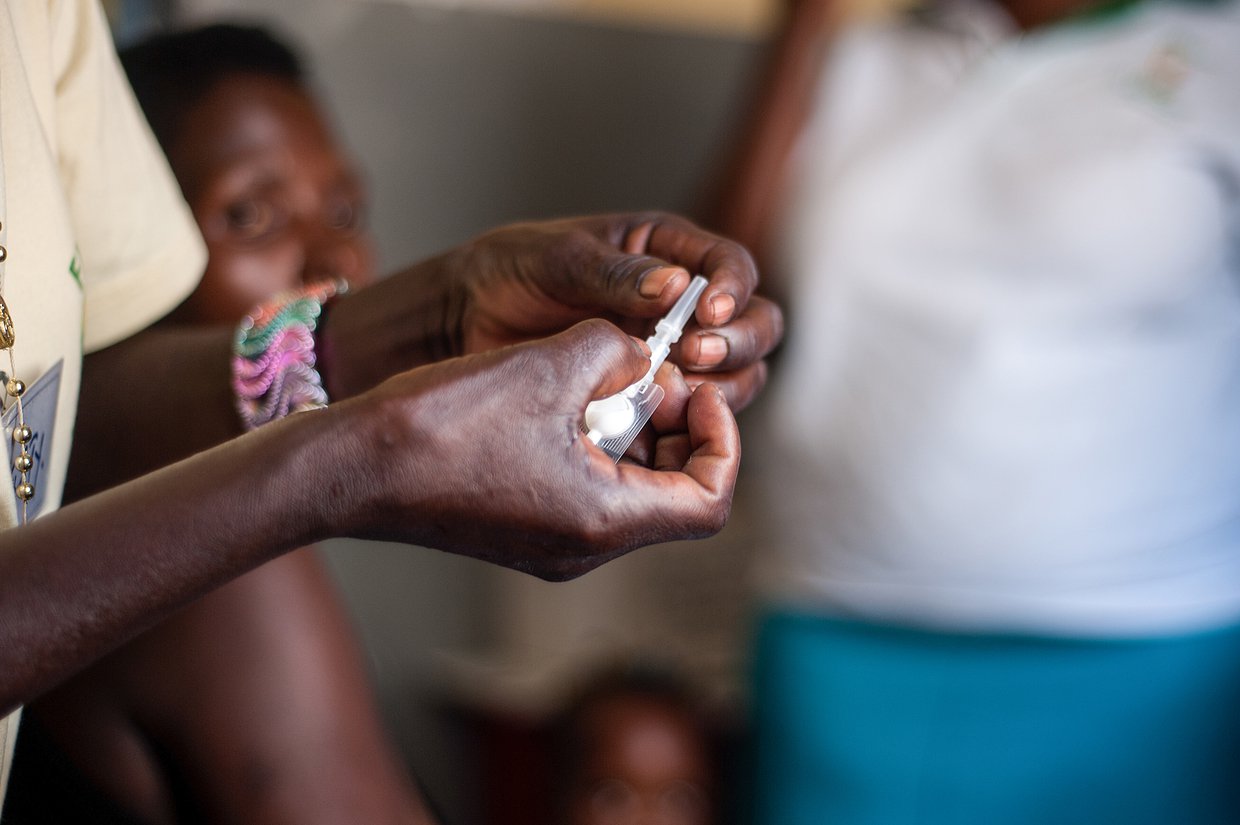 A close-up of a health worker's hands holding a Sayana Press device, with other women in the background.