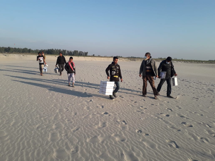 Vaccinators, volunteers, and EPI staff travel by boat and carry vaccines by foot on a river island in Kurigram District. Because of the scattered island geography in the Brahmaputra River and lack of roads, transport by foot is often the only option.