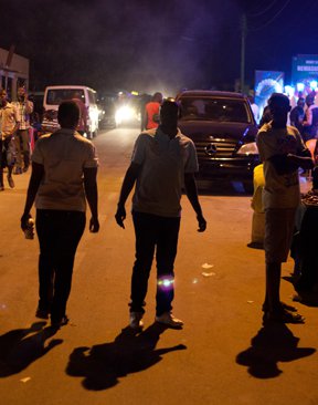 People backlit by headlights stroll down a city street at night.