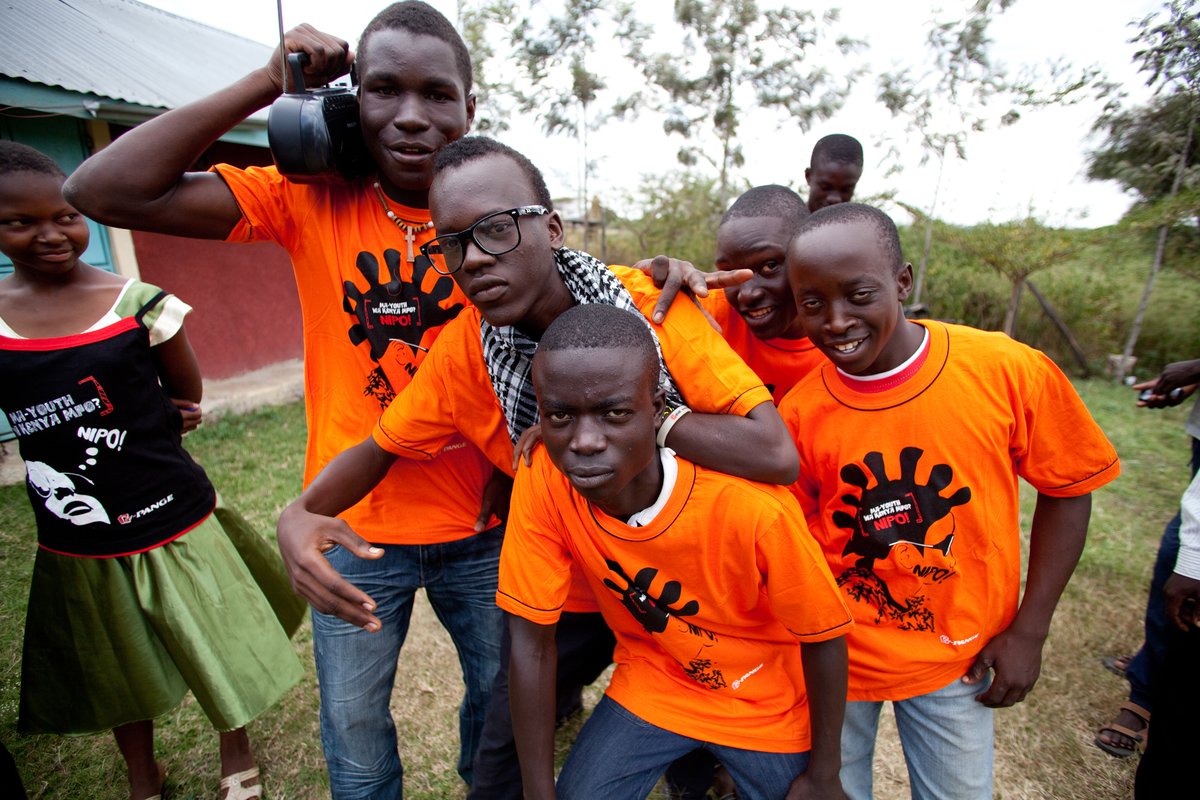A young woman watches five young men in bright orange T-shirts mug for the camera, one carrying a portable radio on his shoulder.