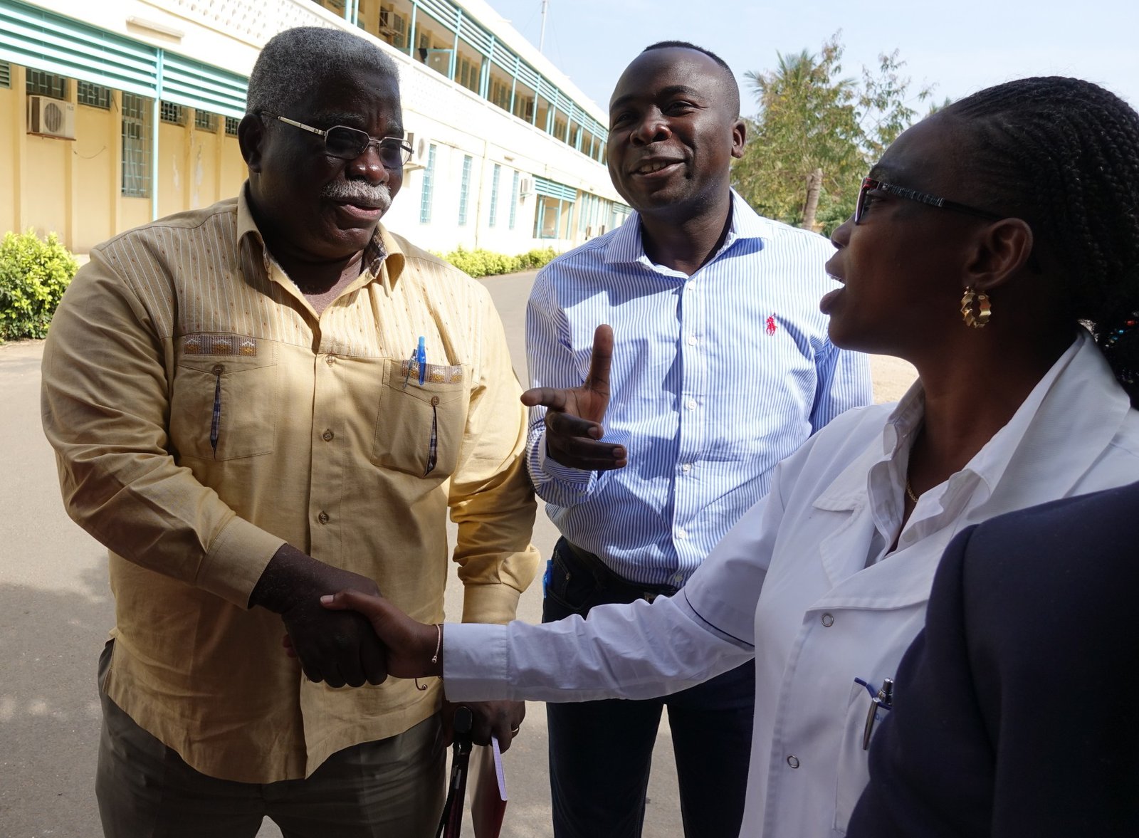 Two people shake hands outside while a third person looks on. 