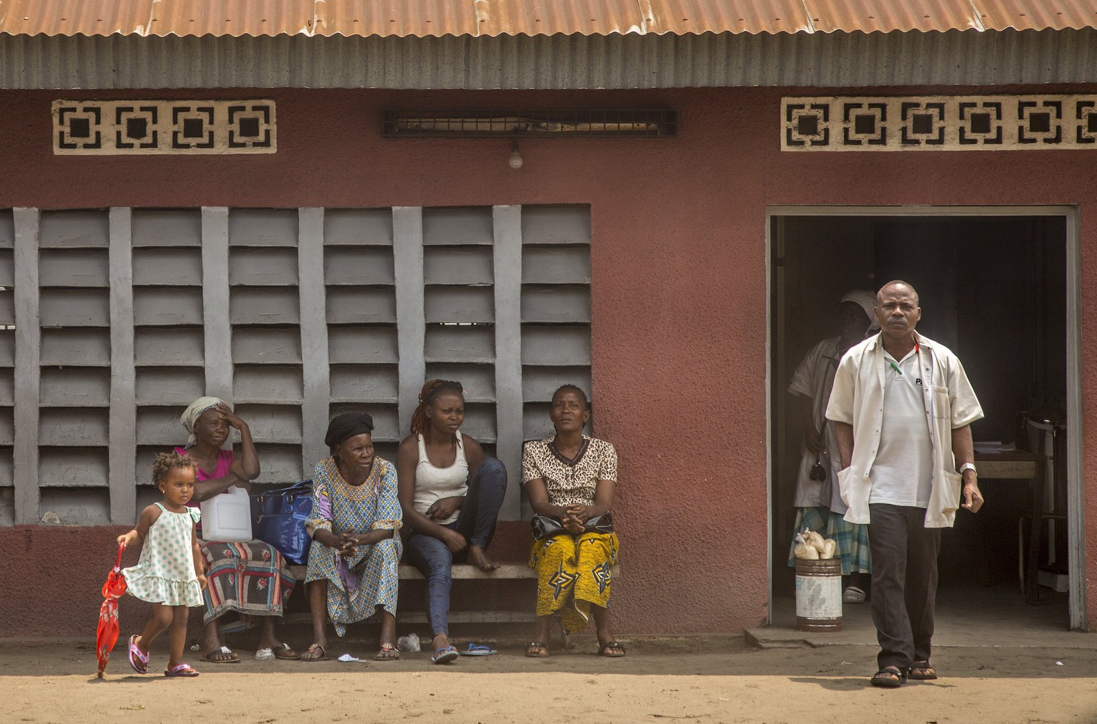 A row of women sit on a bench in front of a clinic. A man stands in the doorway.