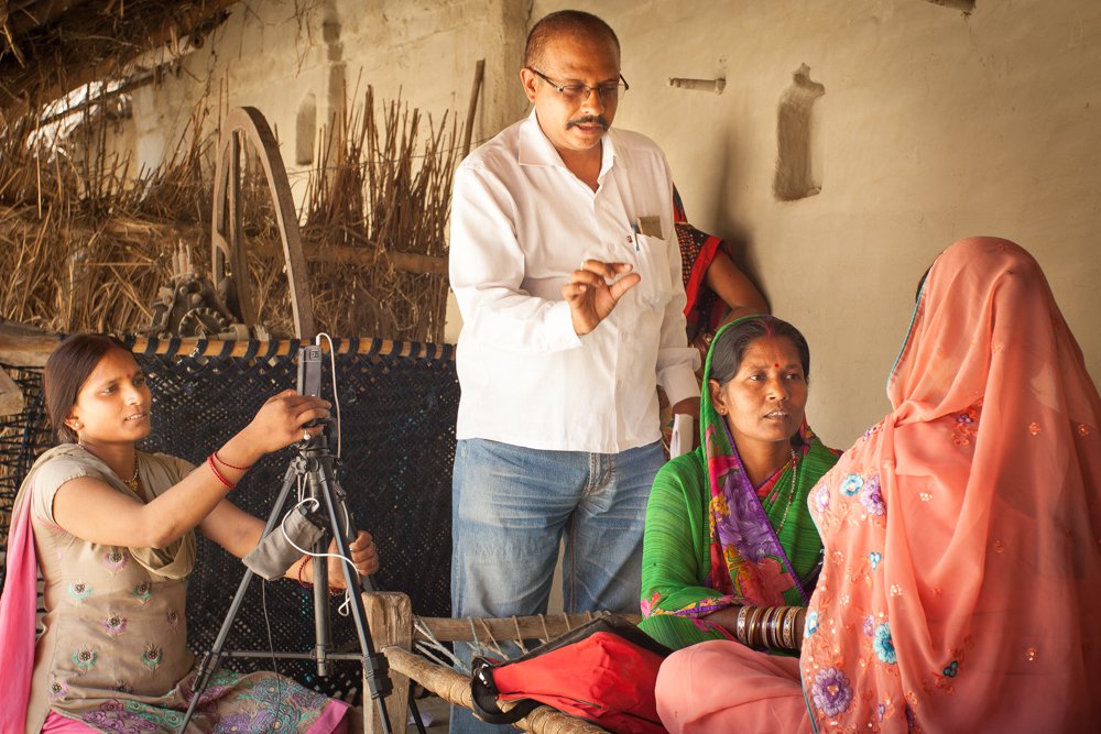 Man directs two women having a conversation as a woman films them with a smartphone on a tripod.