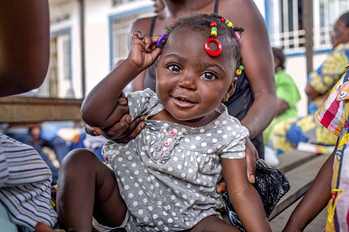 A female toddler with bright hair ties and in a polka-dot jumper is held by a woman.