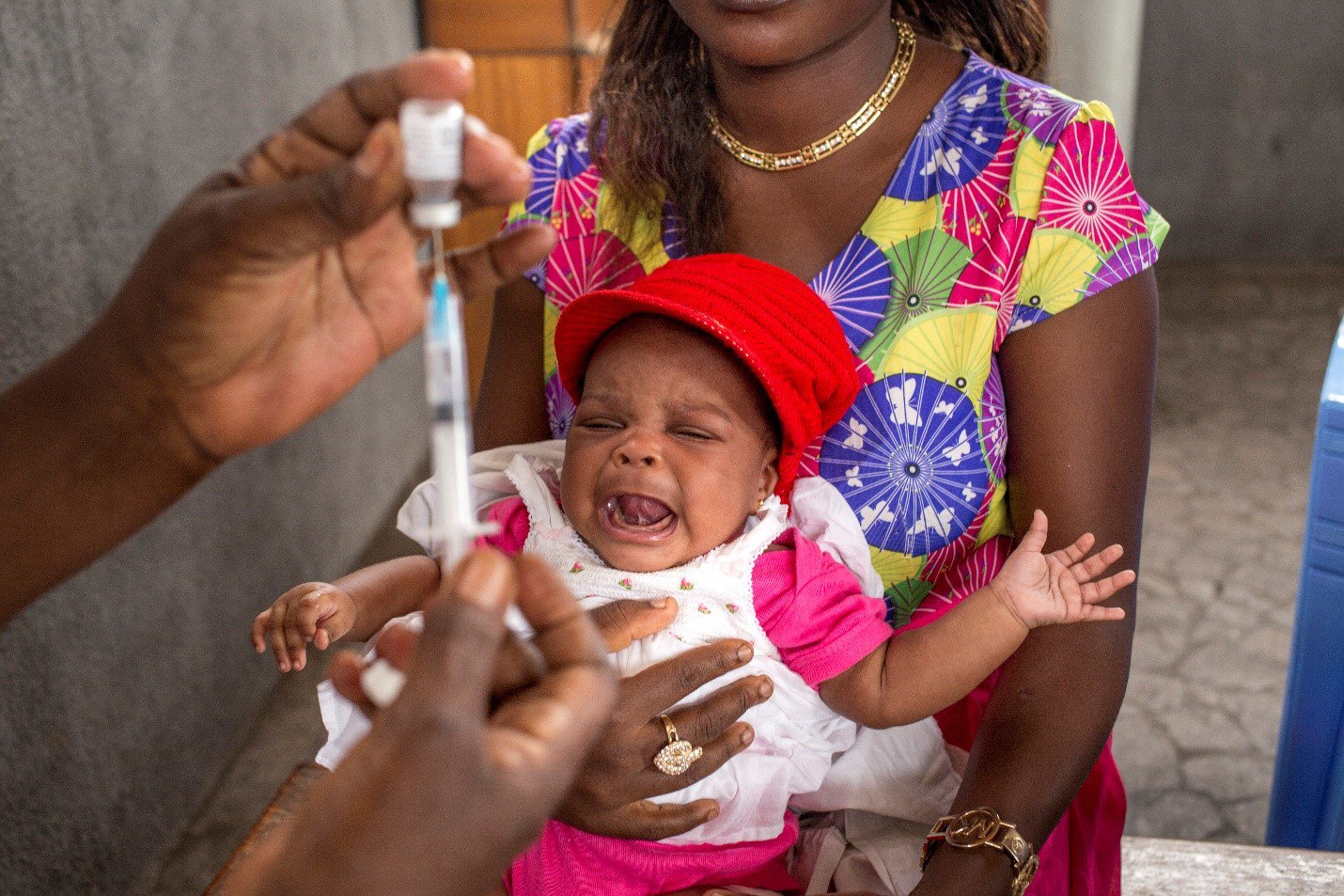 A young child is held on a woman's lap as a healthworker (in the foreground) prepares a vaccine.