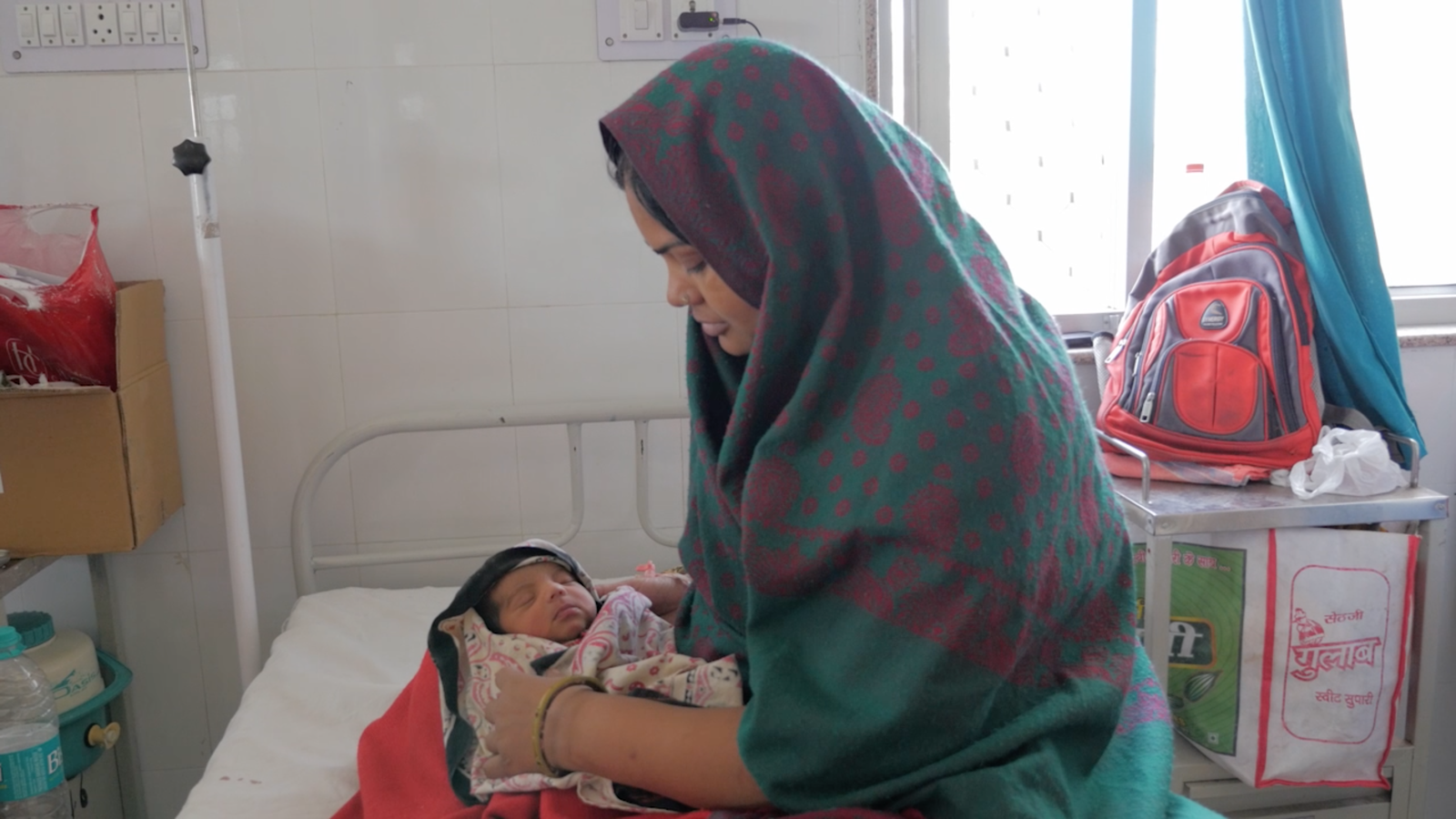 A woman sits on a nursery bed and looks down upon a newborn who is swaddled in a blanket.