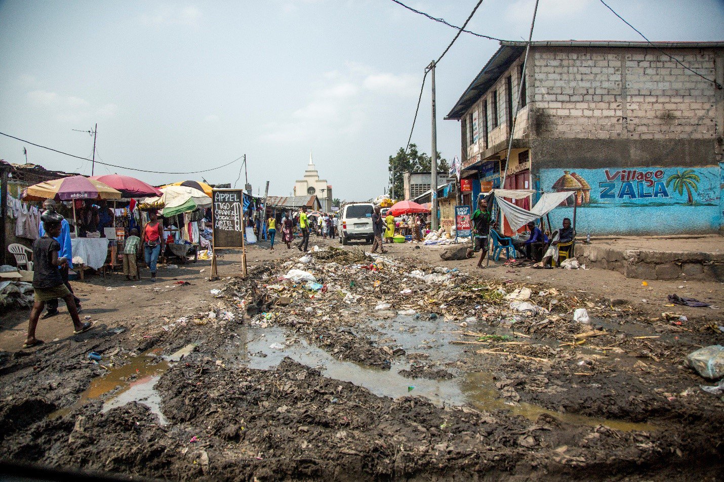 A mud-rutted street is lined with people, buildings, and colorful umbrellas.