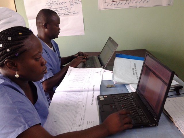 Two people in scrubs working on laptops and a large printed spreadsheet.