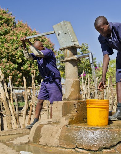 One young man works the handle of a water pump while another stations an orange bucket beneath the spigot.