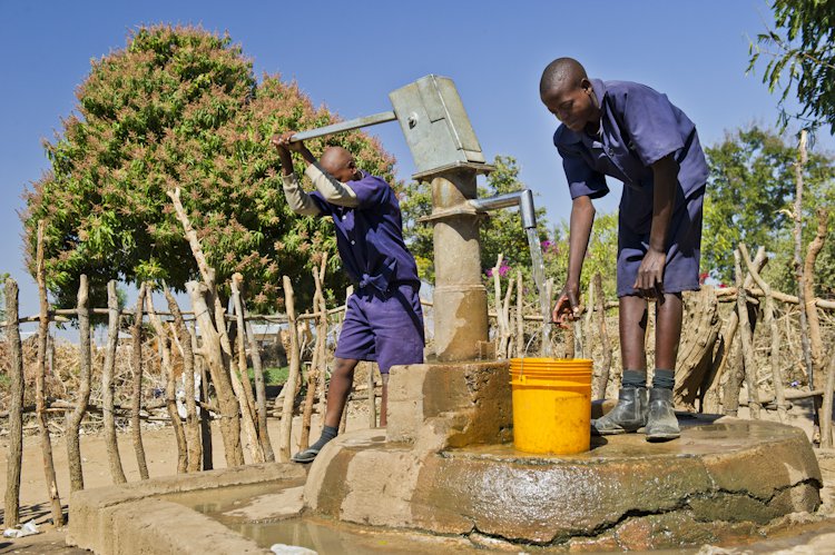 One young man works the handle of a water pump while another stations an orange bucket beneath the spigot.